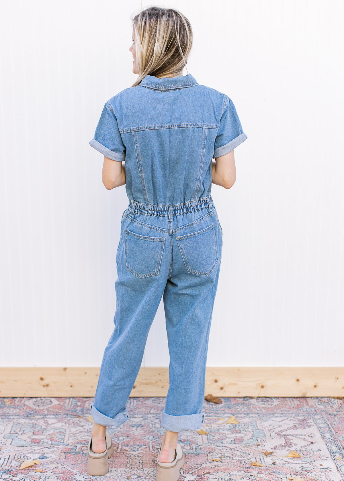 Back view of model wearing a medium washed, short sleeve denim jumpsuit with an elastic waist.