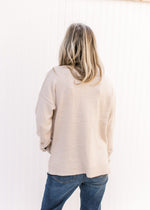 Back view of a model wearing a taupe sweater with floral detail on long sleeves and split sides. 