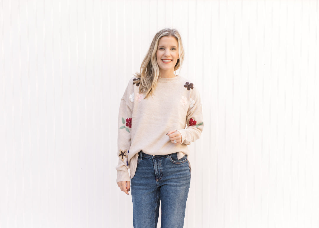 Model wearing a taupe sweater with a floral detail on the sleeves, a round neck and split sides. 