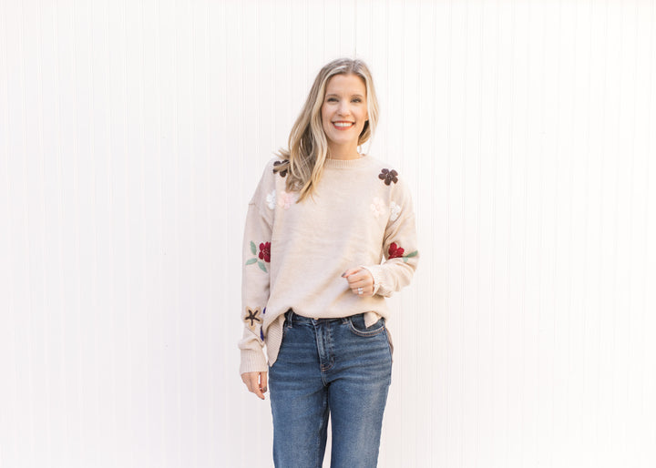 Model wearing a taupe sweater with a floral detail on the sleeves, a round neck and split sides. 