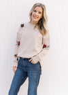 Model wearing jeans with a taupe sweater with floral detail on long sleeves and a round neck.