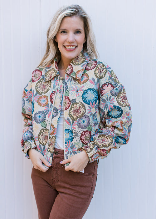 Model wearing a cream zip-up jacket with colorfully embroidered flowers and long sleeves. 