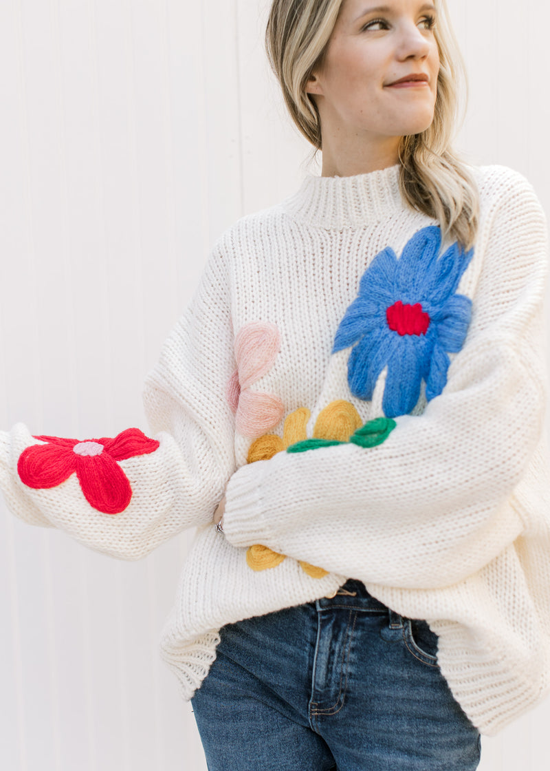 Model wearing jeans and a cream oversized knit sweater with red, yellow, blue, pink and red flowers.