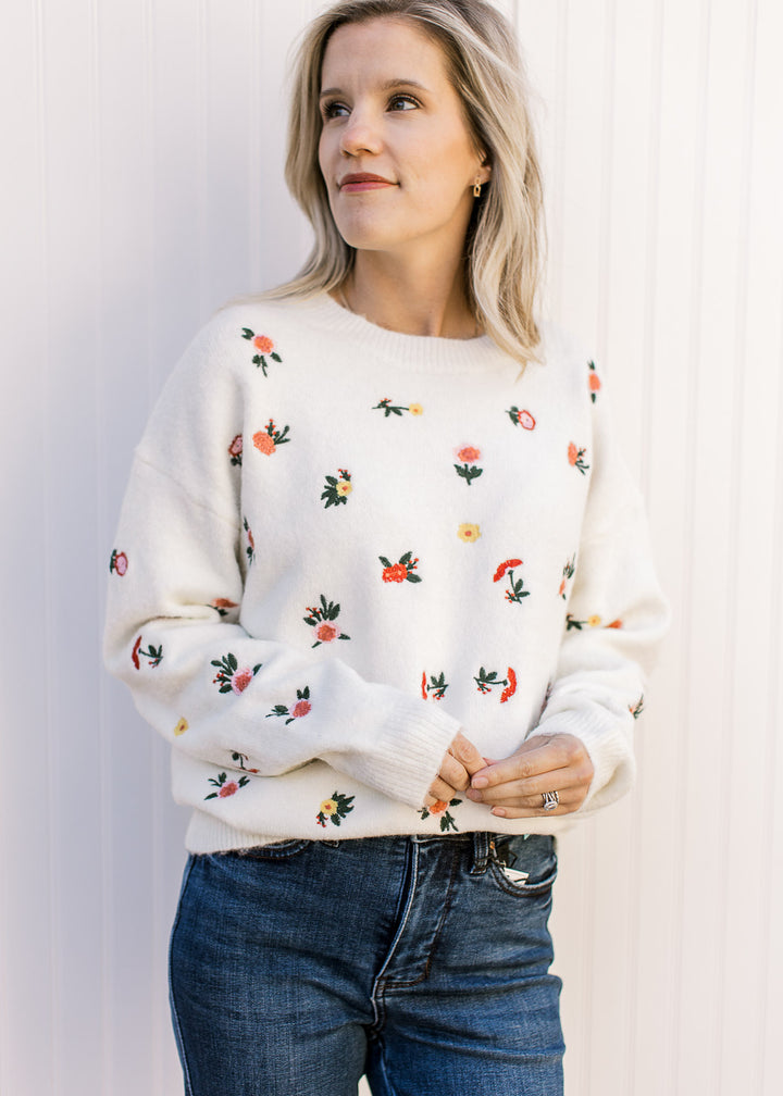 Model wearing a cream sweater with pink and yellow flowers, round neck and long sleeves. 