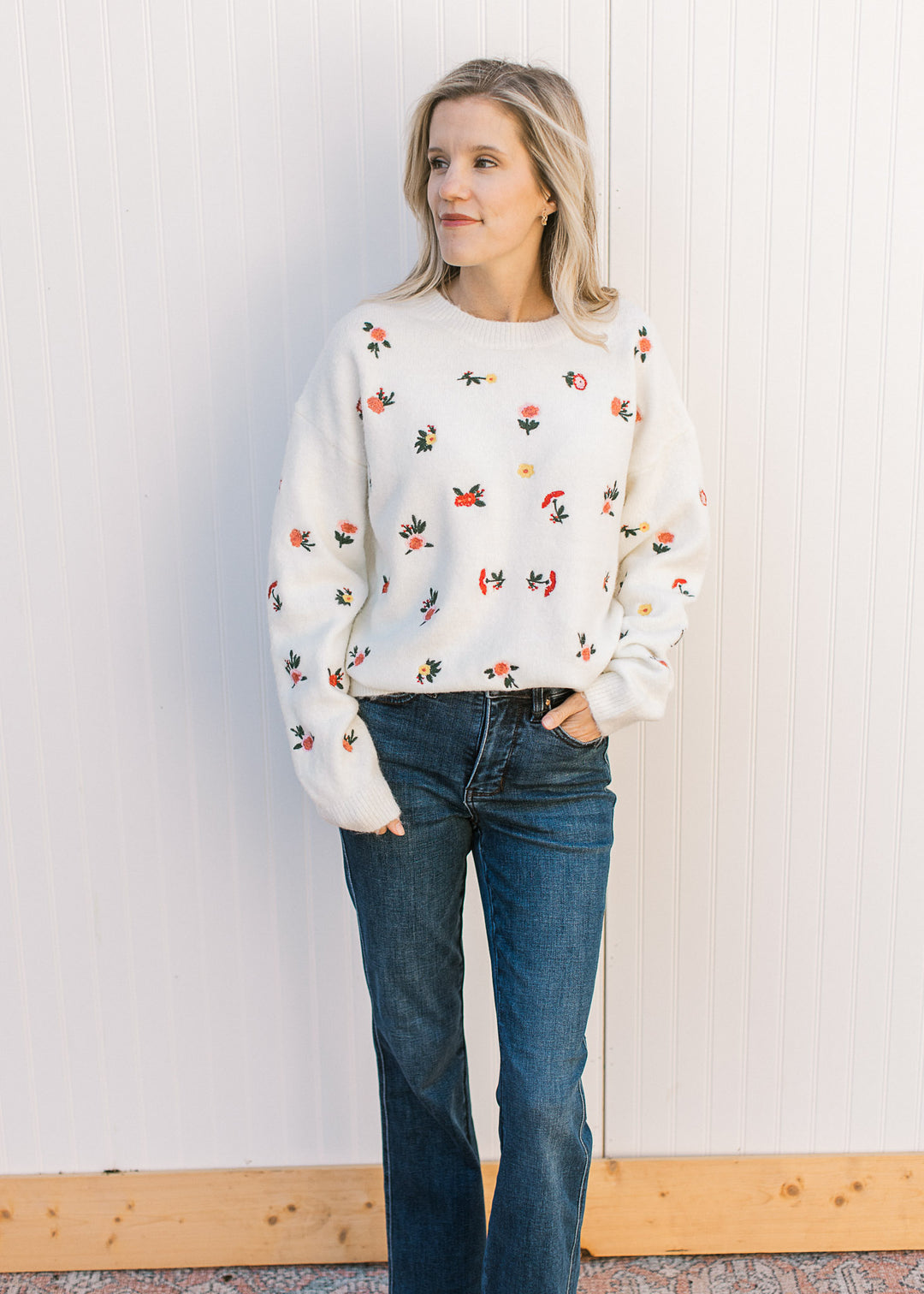 Model wearing jeans and a cream sweater with pink and yellow flowers, round neck and long sleeves. 