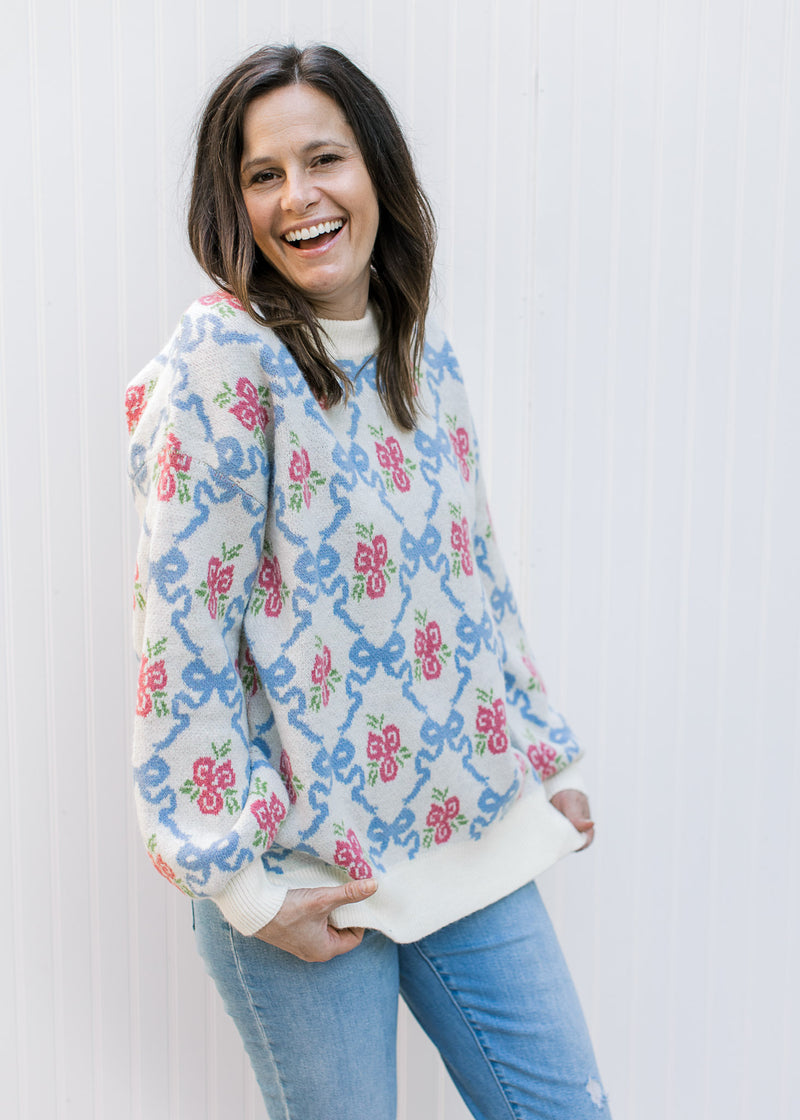 Model wearing jeans and a cream top with blue bows and pink flowers, long sleeves and round neck.