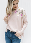 Model wearing a blush sweater with pink, cream, green and white embroidered flowers and a mock neck.