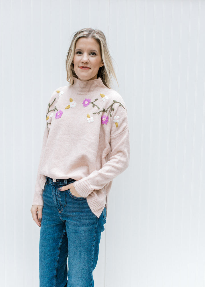 Model wearing jeans and a blush sweater with embroidered flowers and a mock neck.