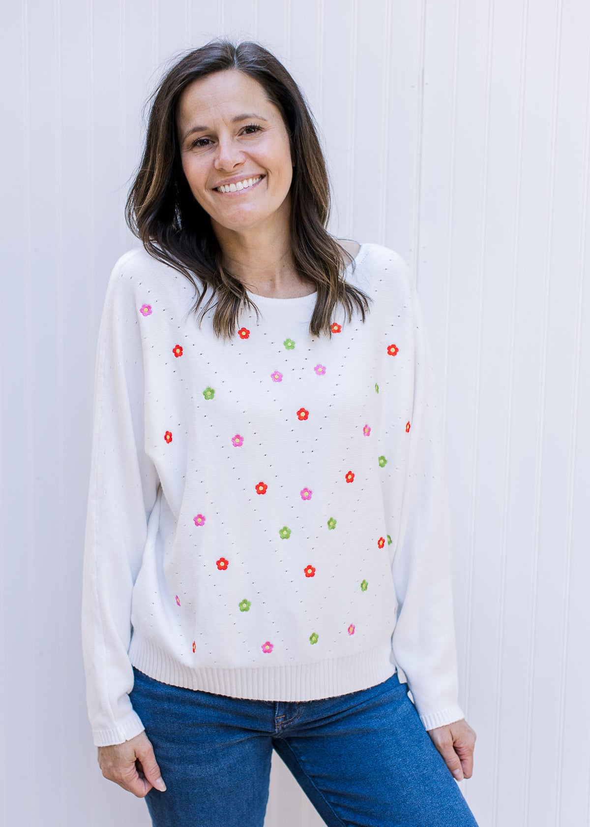 Model wearing jeans and a white sweater with colorful embroidered flowers and long sleeves. 