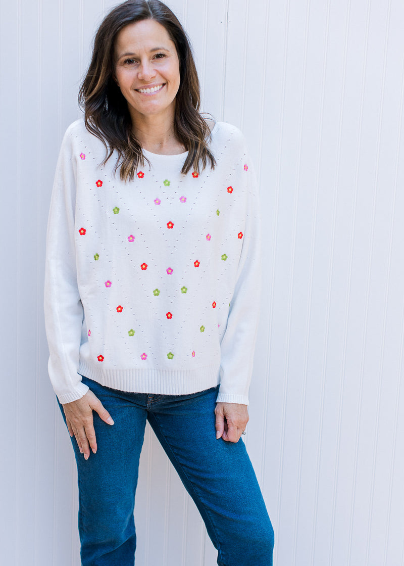 Model wearing a white long sleeve sweater with colorful embroidered flowers. 