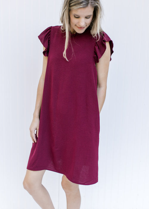 Model wearing a wine above the knee dress with a ruffle cap sleeve and a round neck with a ruffle. 