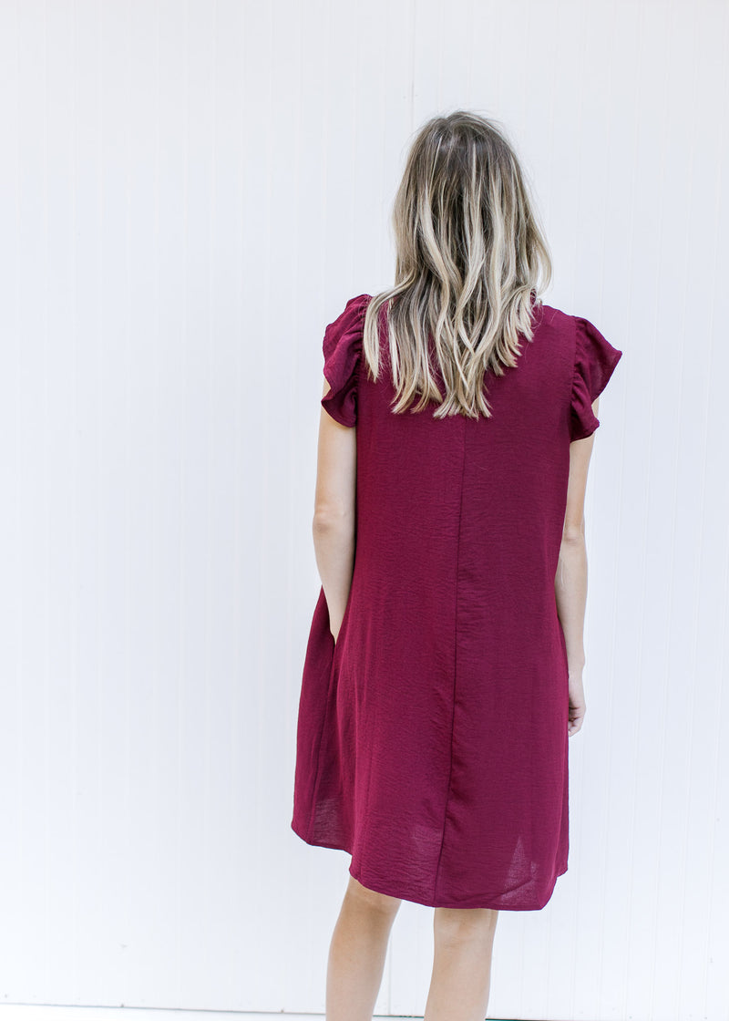 Back view of a Model wearing a wine above the knee dress with a ruffle at the neck and  cap sleeve. 