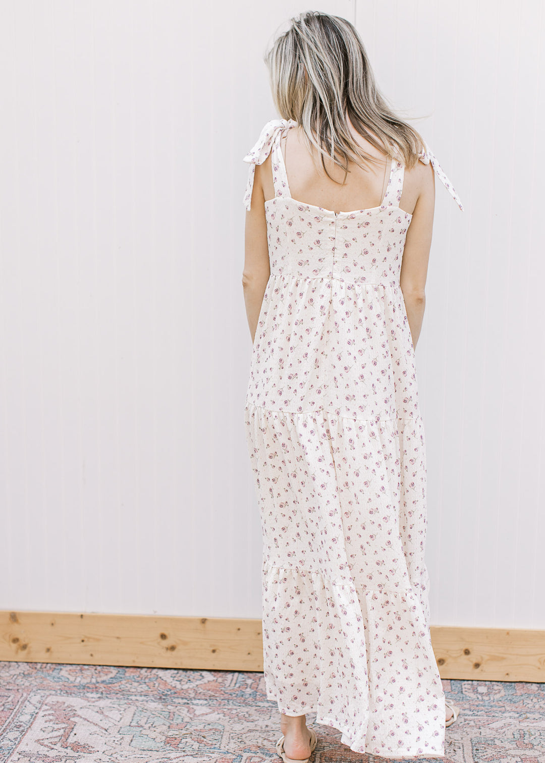 Back view of model wearing a cream midi with purple floral, v-neck, tie straps and tiered skirt.