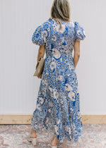 Back view of Model wearing a blue maxi dress with cream floral, ruffle neck and short puff sleeves.