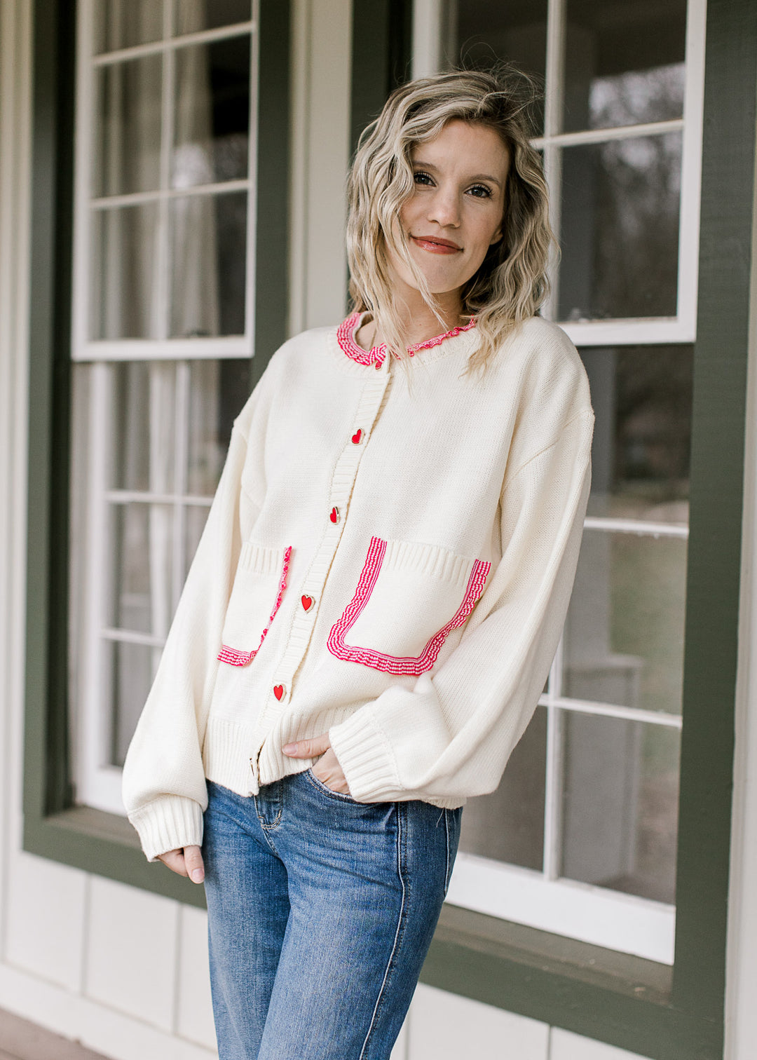 Model wearing a cream cardigan with ruffles at neck and pockets, heart buttons and long sleeves.