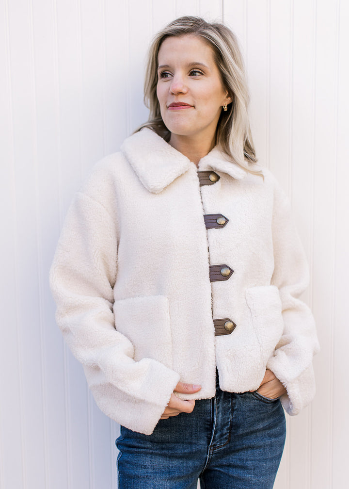 Model wearing a fuzzy cream jacket with faux leather strap with gold buttons and patch pockets.