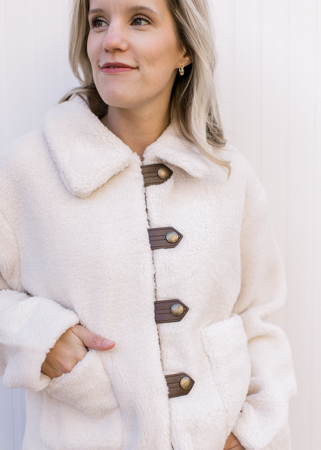 Close up of faux leather straps with gold buttons on a fuzzy cream jacket with patch pockets. 
