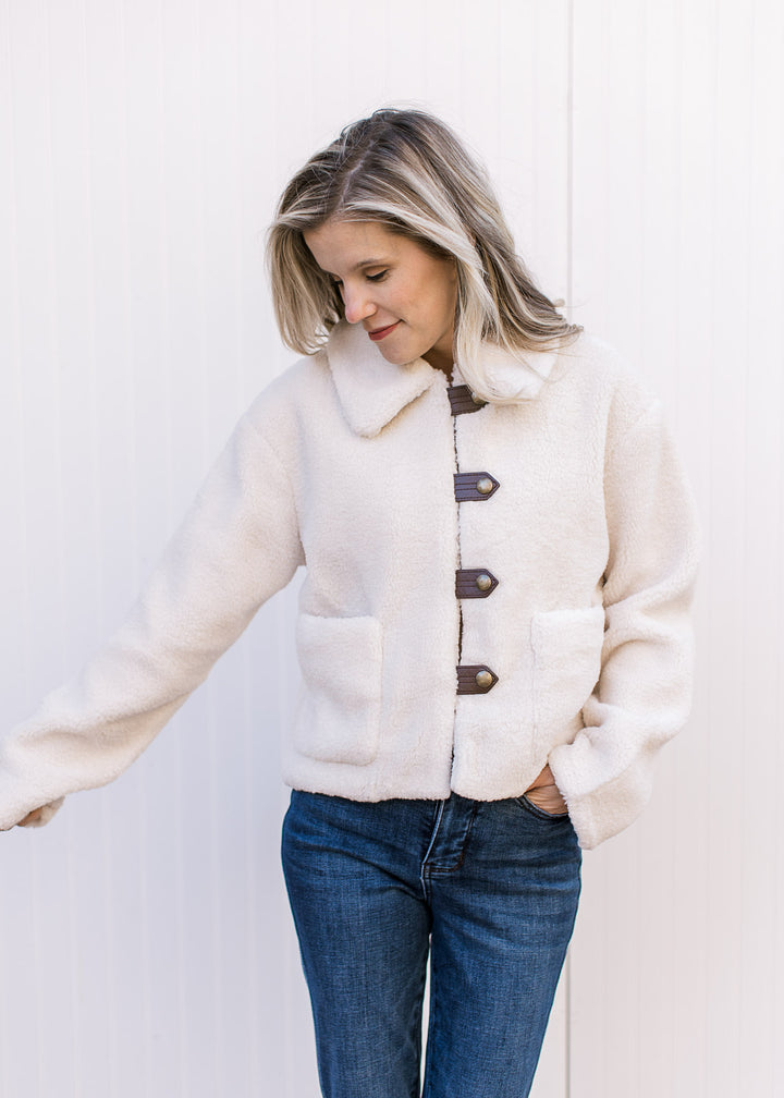 Model wearing a fuzzy cream jacket with faux leather strap, long sleeves and patch pockets.