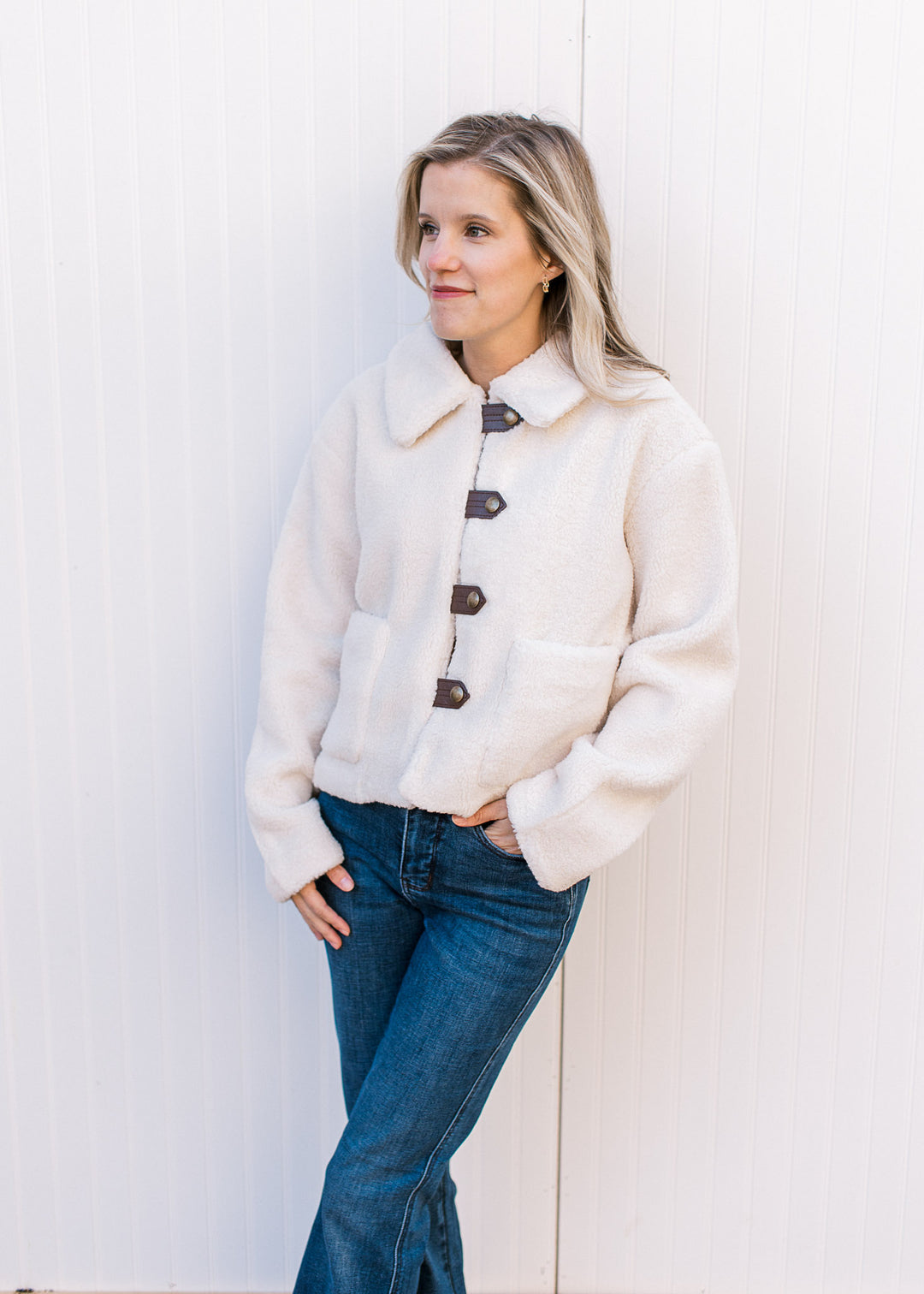 Model wearing jeans and a fuzzy cream jacket with faux leather strap with buttons and patch pockets.