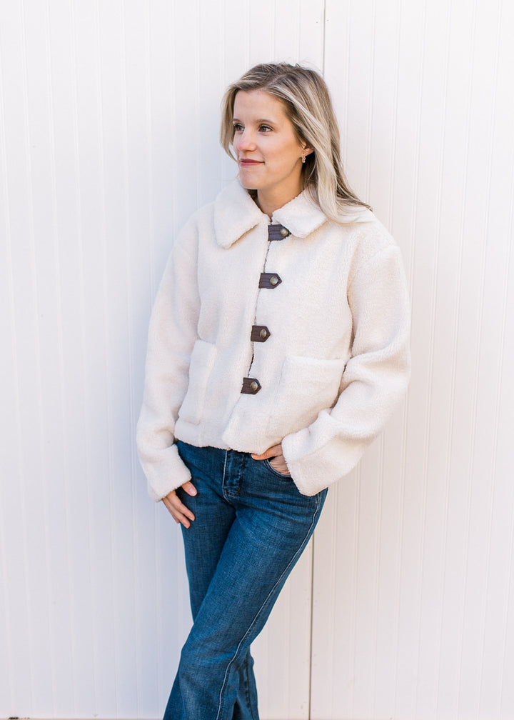Model wearing jeans and a fuzzy cream jacket with faux leather strap with buttons and patch pockets.