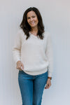 Model wearing jeans with a cream long sleeve fuzzy sweater with pearl embellishments at v-neck. 