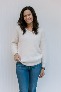 Model wearing jeans with a cream long sleeve fuzzy sweater with pearl embellishments at v-neck. 