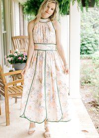 Model wearing heals and a pale pink midi with a floral design, green piping detail, a halter fit.