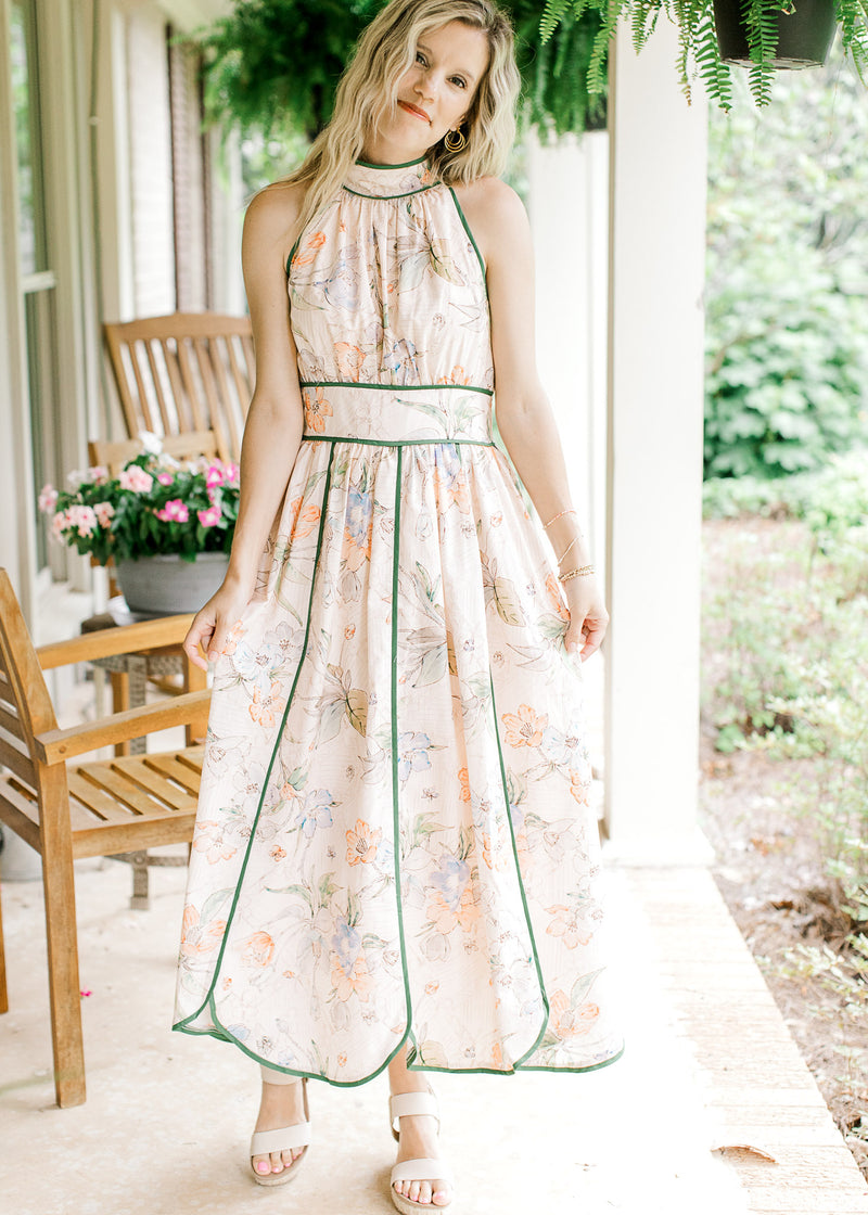 Model wearing heals and a pale pink midi with a floral design, green piping detail, a halter fit.