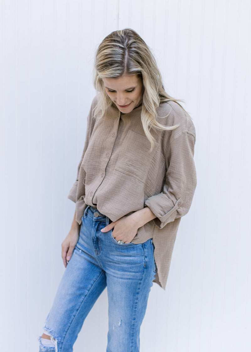 Model wearing a taupe button up top with a raw hemline, a cotton gauze material and long sleeves. 