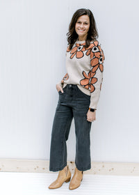 Model wearing black jeans with a taupe sweater with a round neck and rust flowers outlined in black.