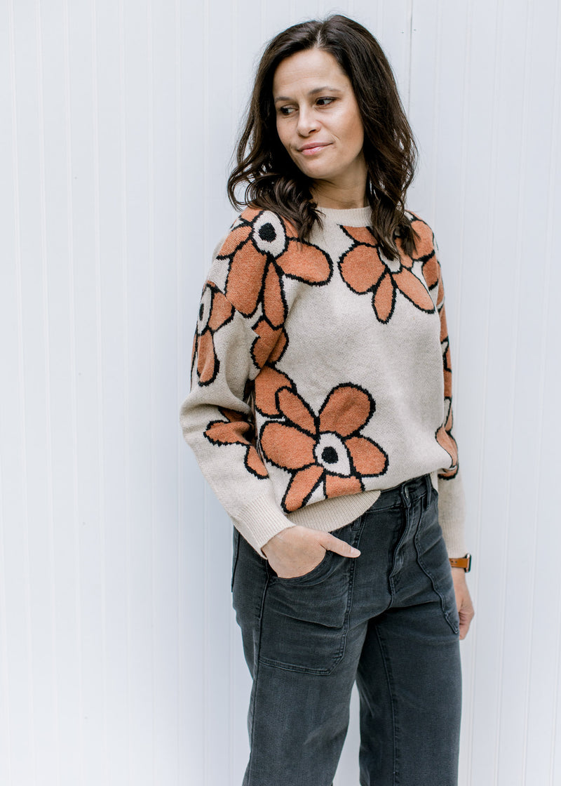 Model wearing a taupe sweater with a round neck, long sleeves and rust flowers outlined in black. 