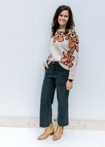 Model wearing jeans, mules and a taupe sweater with a round neck and rust flowers outlined in black.