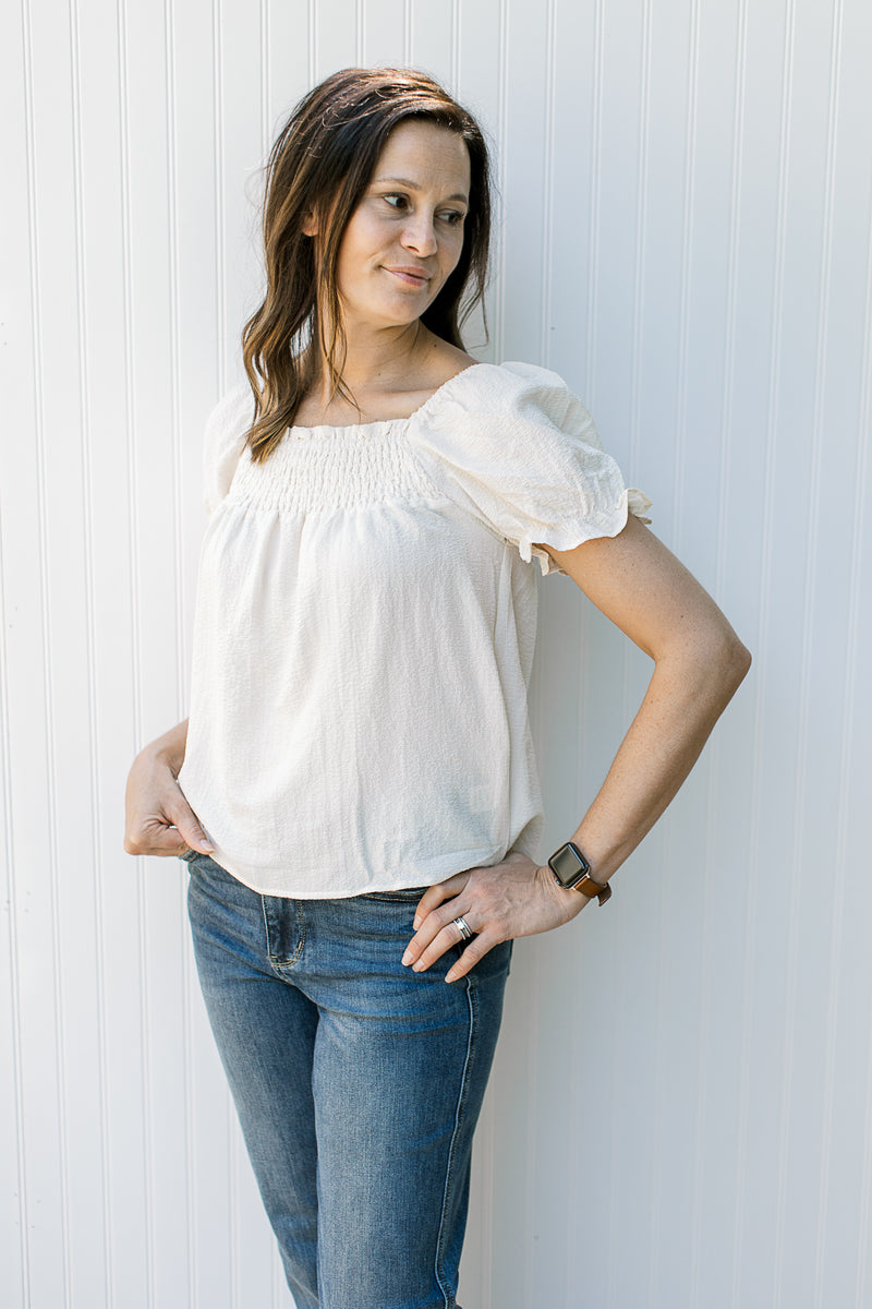 Model wearing jeans with a cream top with a smocked yolk, bubble short sleeves and a square neck.