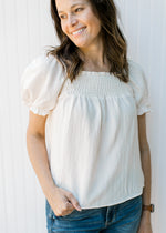 Model wearing a cream top with a smocked yolk, bubble short sleeves and a square neck.