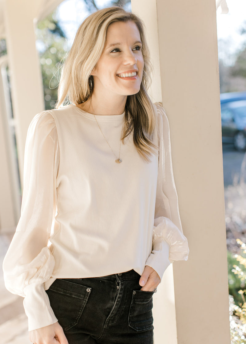 Model wearing jeans and an ivory top with shimmer long sleeves and a ribbed bodice. 