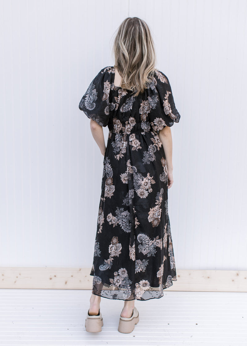 Back view of Model wearing a black maxi with tan and gray flowers, short sleeves and a square neck.
