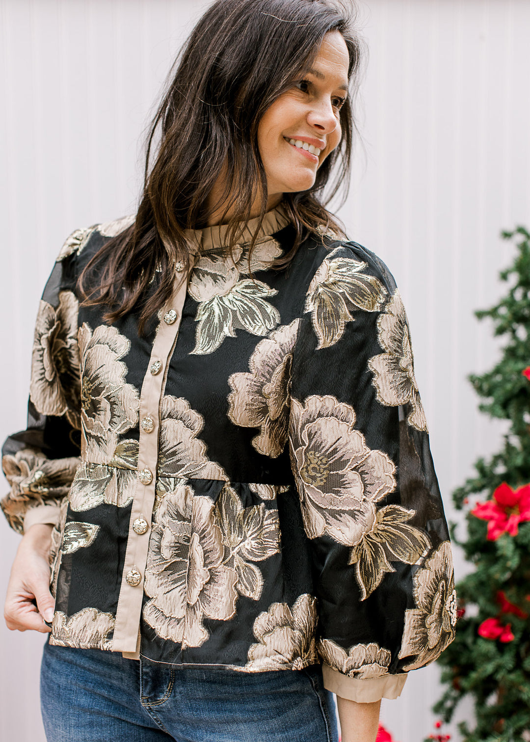 Model wearing a black top with gold floral, button front with taupe details and a ruffle mock neck. 