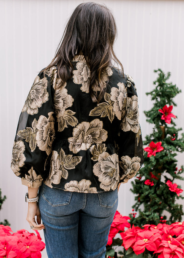 Back view of Model wearing a black top with gold floral and taupe details and a ruffle mock neck. 