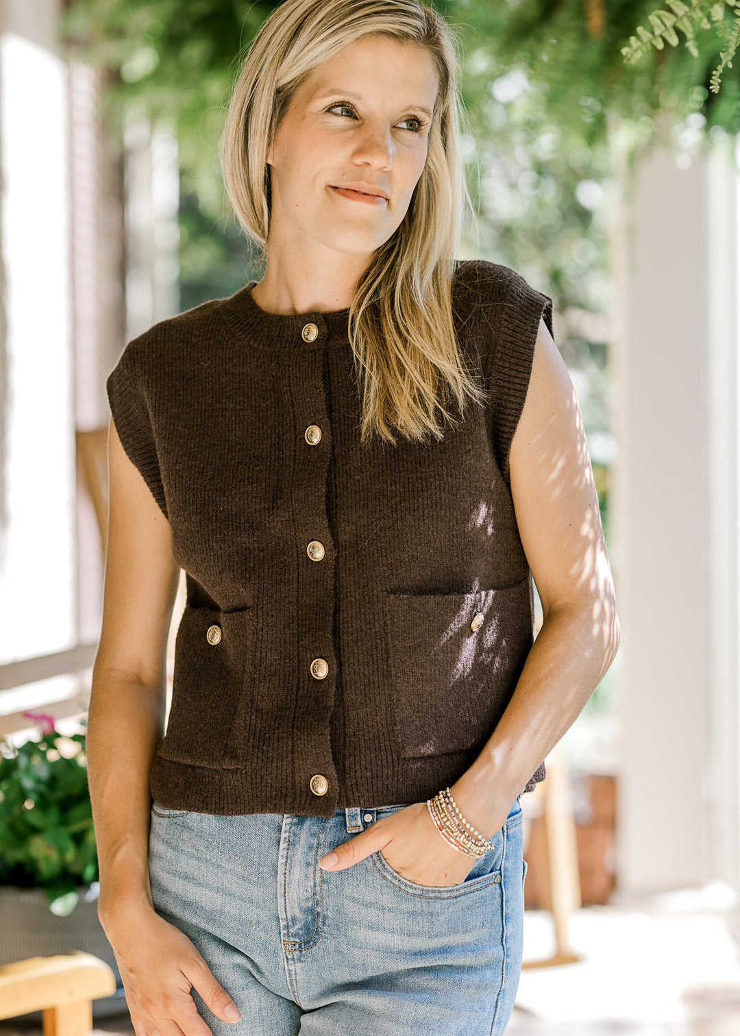 Model wearing a mocha sleeveless top with patch pockets and gold button front. 