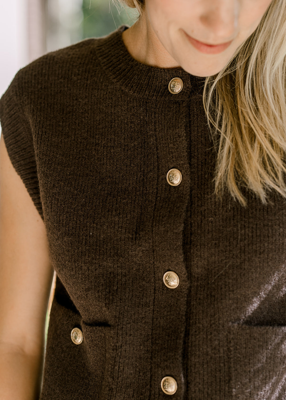 Close up of ribbed detail at neck and cuff of a mocha sleeveless sweater with gold buttons. 