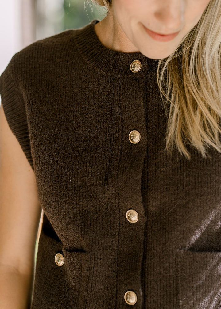 Close up of ribbed detail at neck and cuff of a mocha sleeveless sweater with gold buttons. 