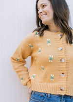 Model wearing a mustard colored button up sweater with yellow and green stitched flowers. 