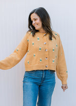 Model wearing jeans with a mustard button up sweater with yellow and green stitched flowers. 