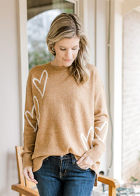 Model wearing jeans and a golden sweater with cream heart pattern, long sleeves and classic styling.