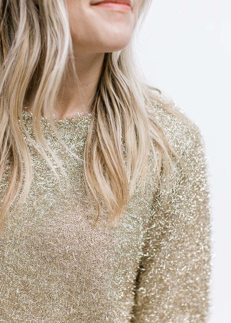 Close up of tinsel material on a gold sweater with a round neck and long sleeves. 