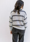 Back view of a Model wearing a cozy gray sweater with black stripes, long sleeves and a round neck. 