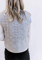 Back view of Model wearing a white top with a gray cable knit vest with a round neck and crop fit. 