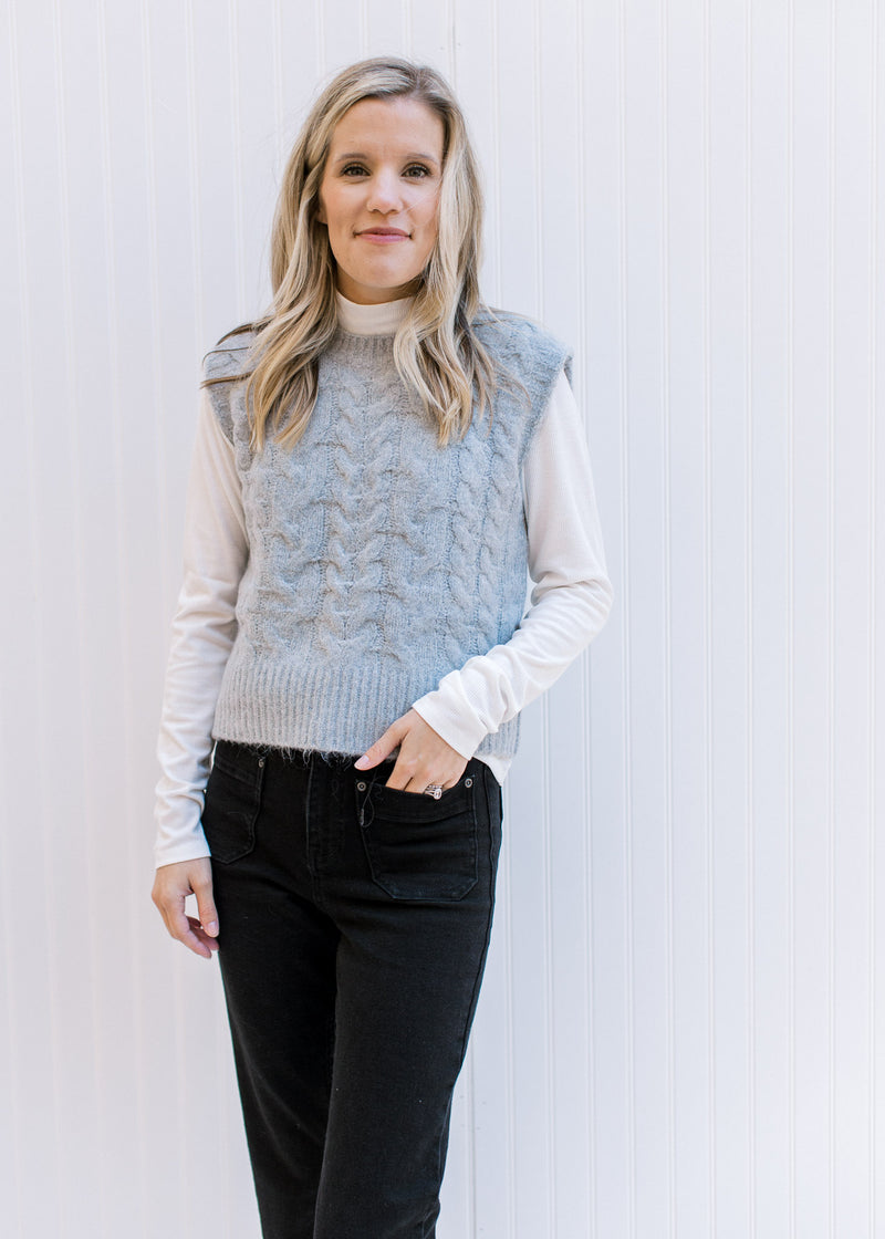 Model wearing a white top, jeans and a gray vest with a round neck and cable knit material. 
