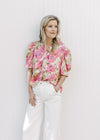 Model wearing a cream short sleeve top with pink and green floral print, buttons and a ruffle neck.