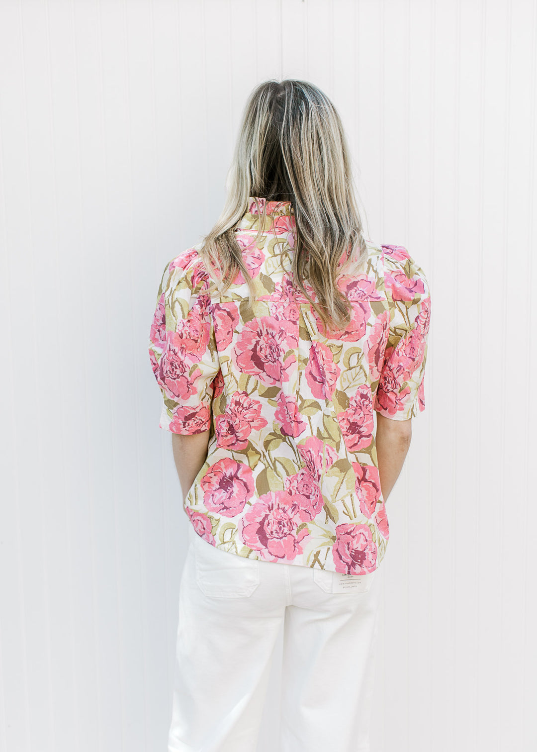 Back view of Model wearing a cream top with pink and green floral print, buttons and a ruffle neck.